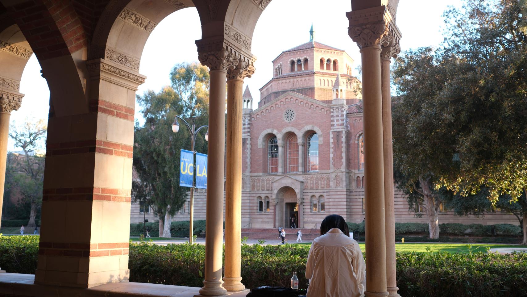 A student sits in an arched opening in the Royce Hall breezeway, facing Powell Library.