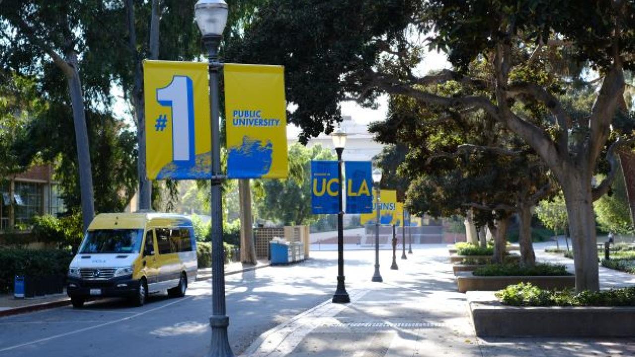 A lamppost-lined street with “#1 Public University” banners, trees, and a parked UCLA van.