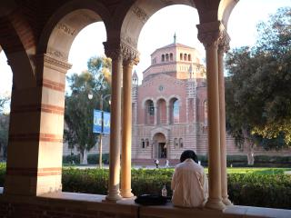 A student sits in an arched opening in the Royce Hall breezeway, facing Powell Library.